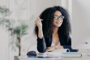 Black woman looks positively at display of computer, writes down information from internet, wears optical glasses and black suit, sits at desktop with necessary things for work to illustrate Marketing During Recessions Time For Bold Moves, Creativity